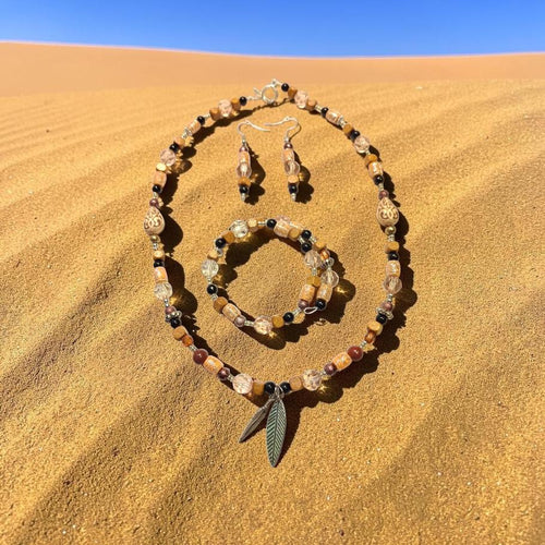 Desert Nomad Jewelry Set displayed on golden sand dunes, featuring carved tan beads, black onyx, quartz crystals and a double feather charm pendant