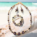 Dreaming in Aloha Jewelry Set on driftwood with turquoise ocean backdrop, featuring hand-painted purple lampwork beads, champagne pearls and silver filigree