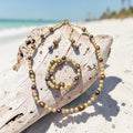 Tropical Elegance Jewelry Set displayed on driftwood at the beach, featuring cream and mauve pearl beads with bronze filigree accents