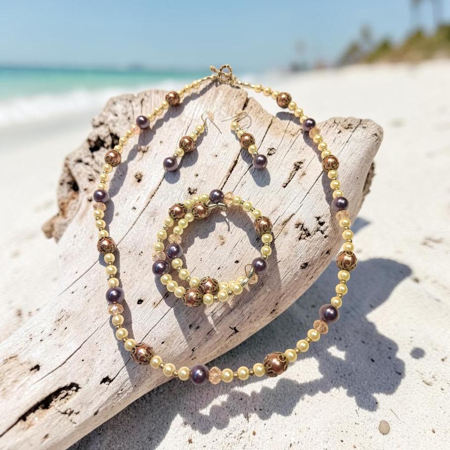 Tropical Elegance Jewelry Set displayed on driftwood at the beach, featuring cream and mauve pearl beads with bronze filigree accents